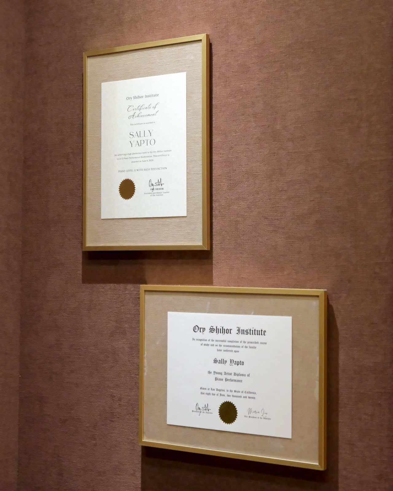Two gold-framed Ory Shihor Institute documents on a rose-coloured wall: a Certificate of Achievement and the Young Artist Diploma of Piano Performance, both awarded to Sally Yapto.