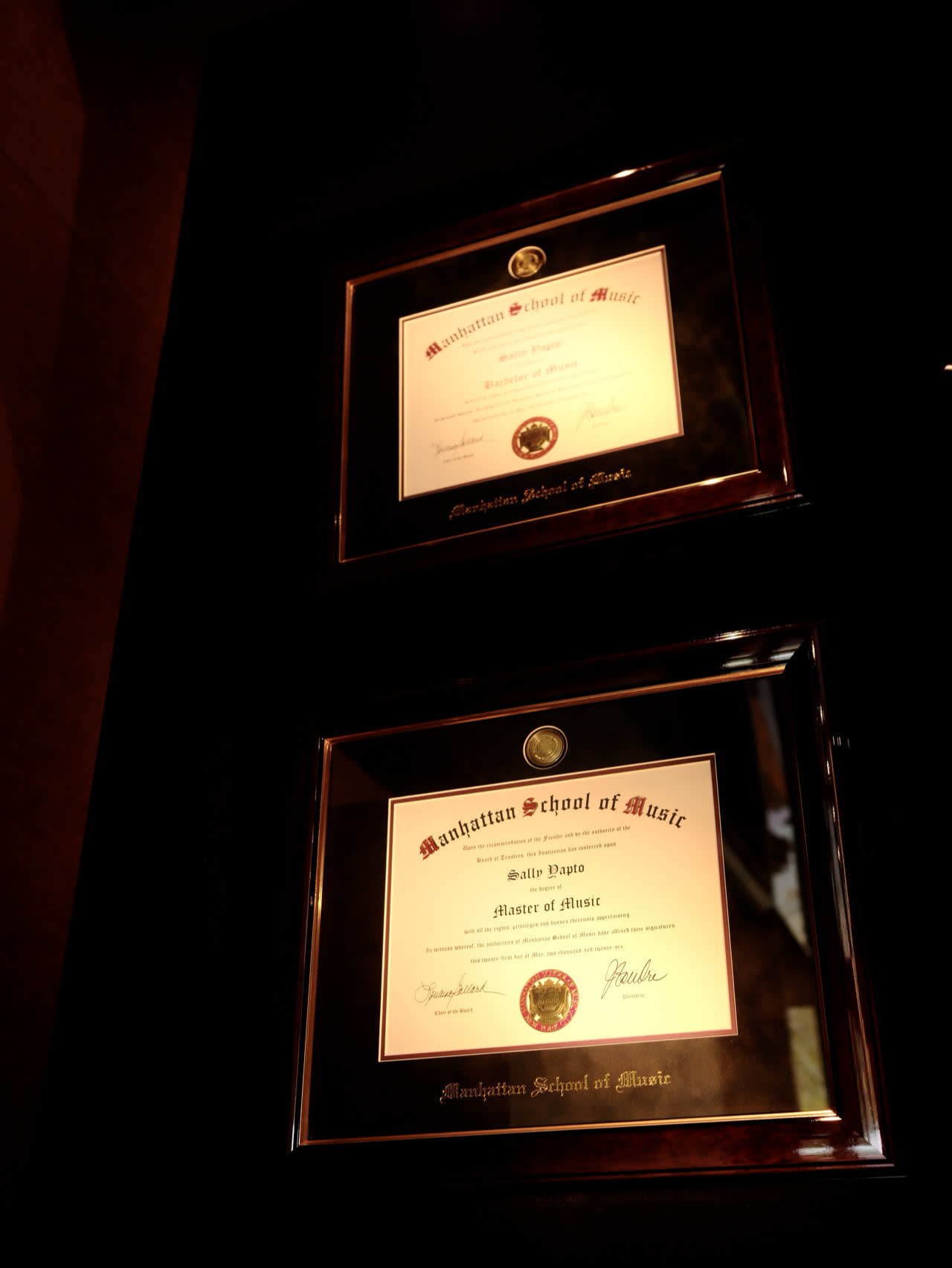 Sally Yapto’s Manhattan School of Music diplomas, Bachelor of Music above, Master of Music below, in dark timber frames on a dark wall.