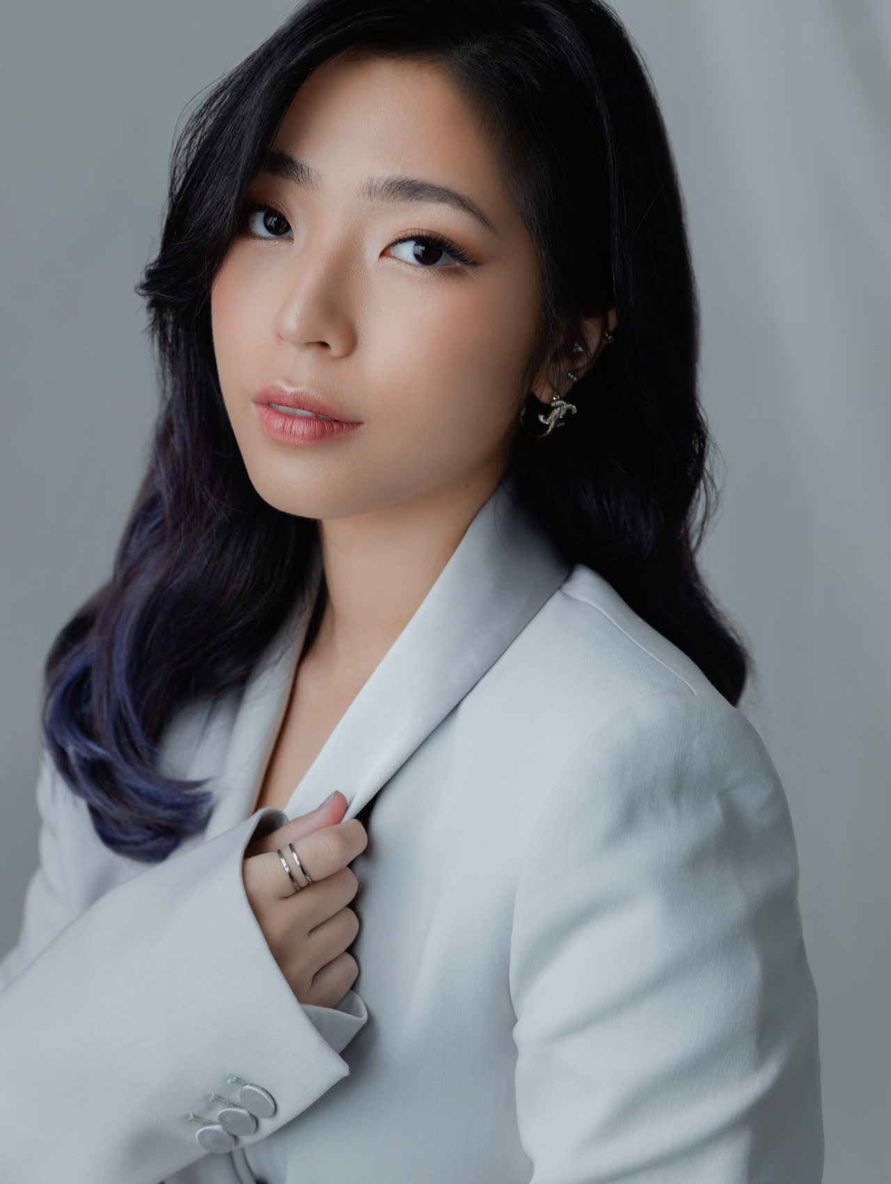 Sally Yapto in a pale grey suit, turned to the camera against a soft grey backdrop.