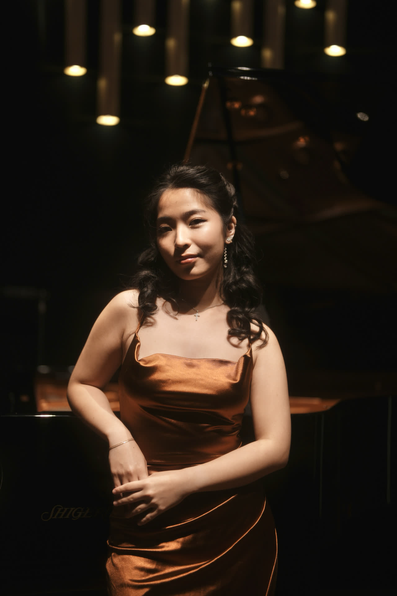 Sally Yapto on stage beside the piano, smiling, in a bronze gown.